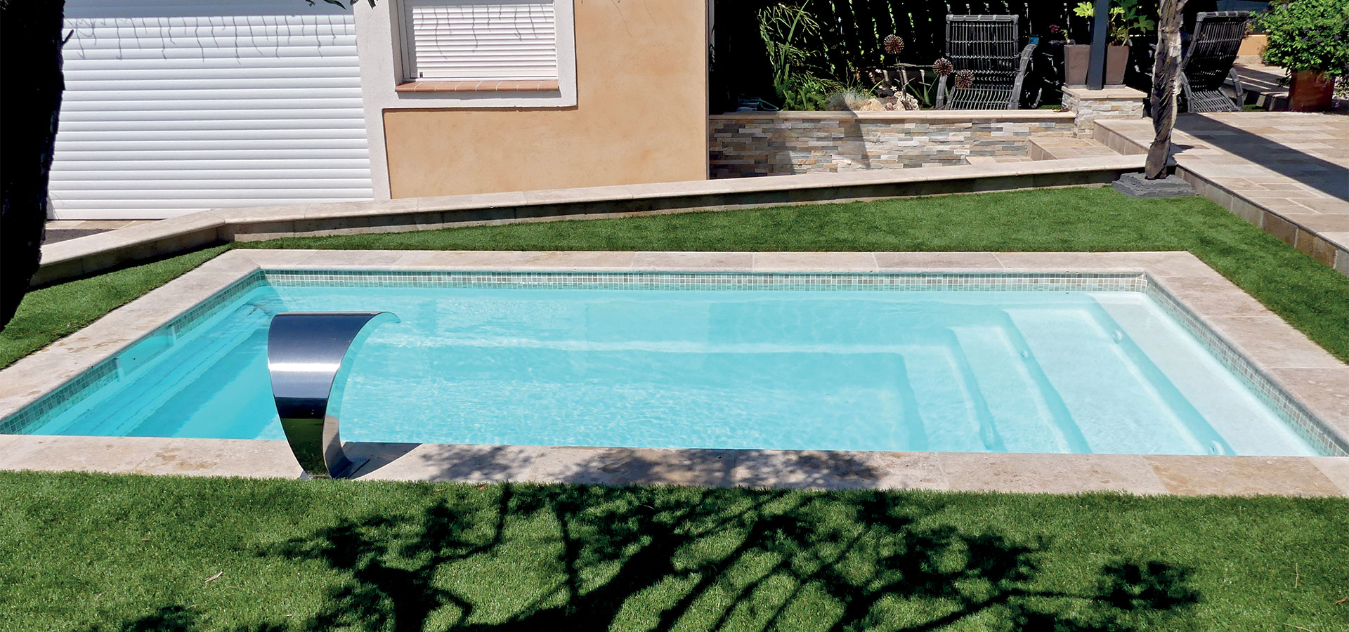 Piscine rectangulaire moderne avec bord en acier inoxydable dans un jardin soigné avec pelouse verte, mobilier de jardin et maisons en arrière-plan.