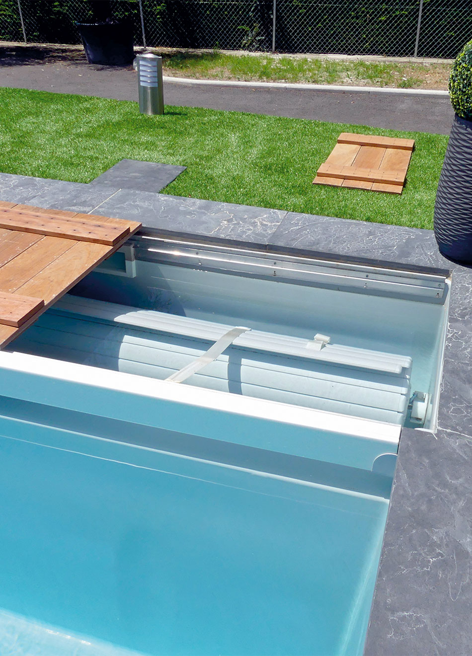 Piscine moderne enterrée avec couverture automatique et terrasse en bois dans un jardin bien entretenu.